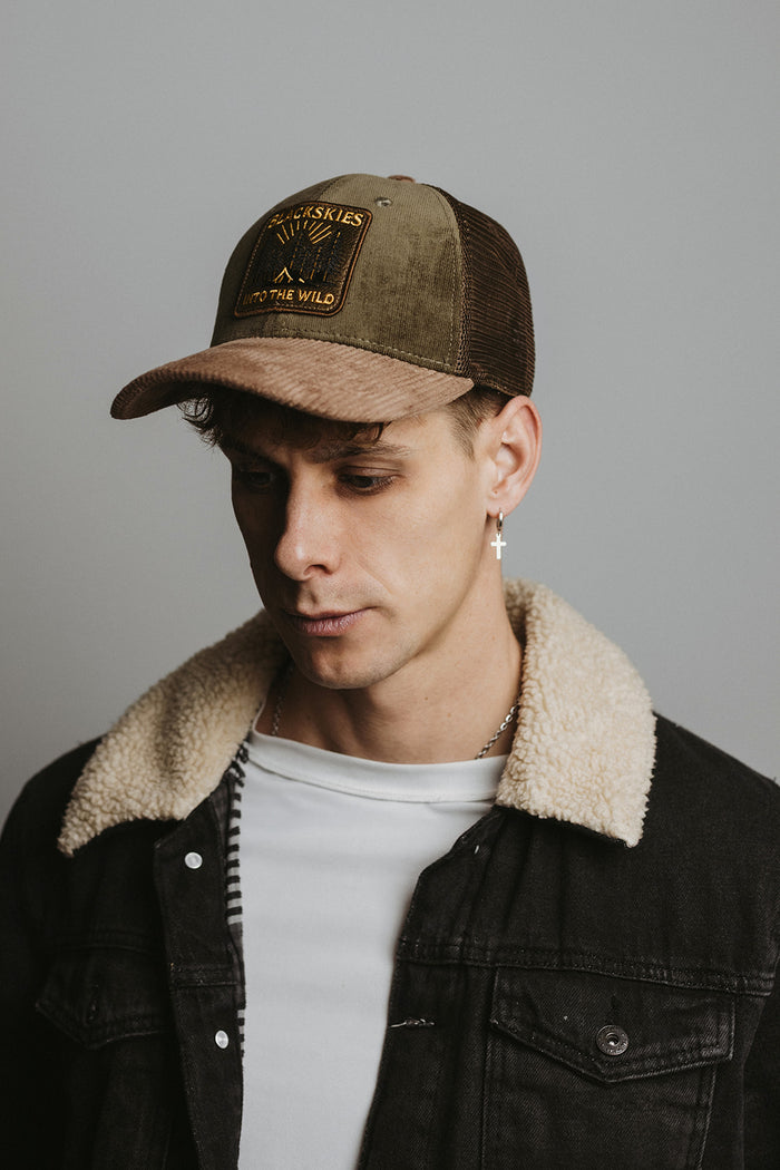 Man wearing a brown cap and black jacket with a shearling collar against a gray background