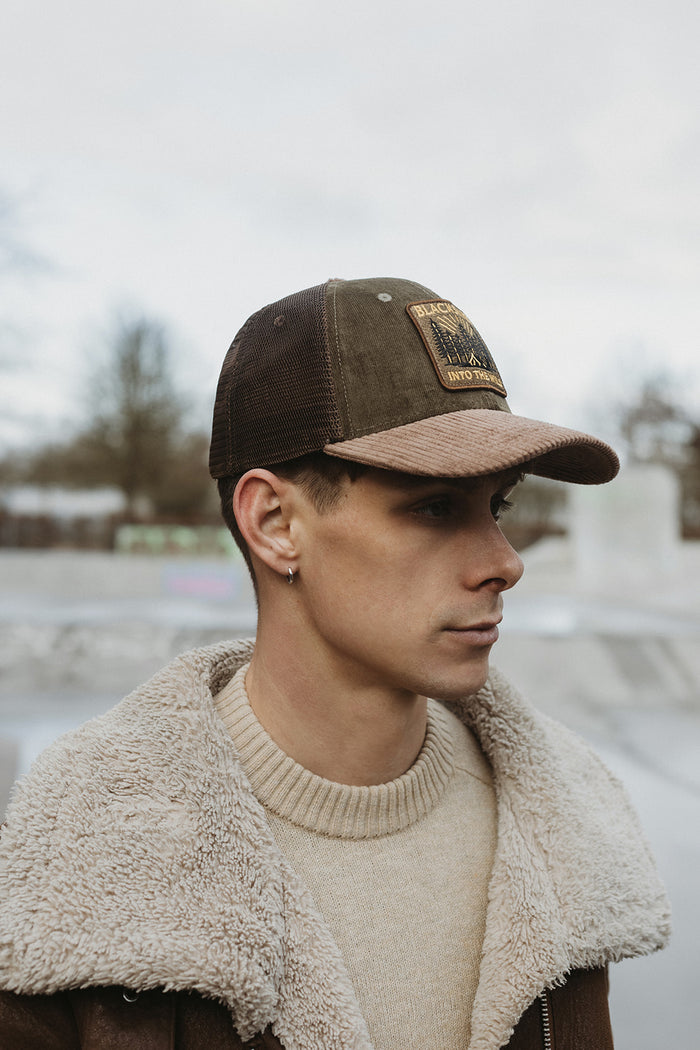 Man wearing a brown cap with a logo and a beige jacket outdoors.