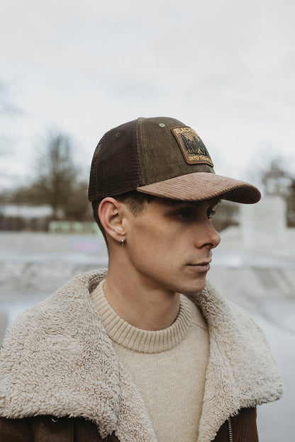 Man wearing a brown cap with a logo and a beige jacket outdoors.