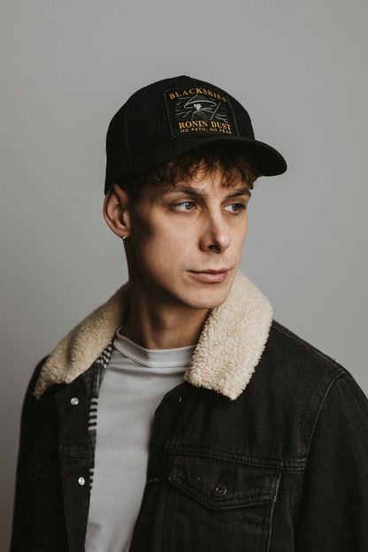 Person wearing a black cap with branding and a dark jacket with a light collar against a plain background