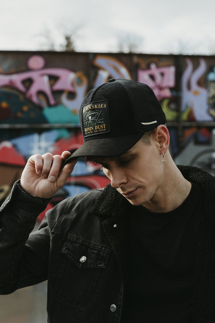 Person wearing a black cap with a logo, standing in front of a graffiti wall.