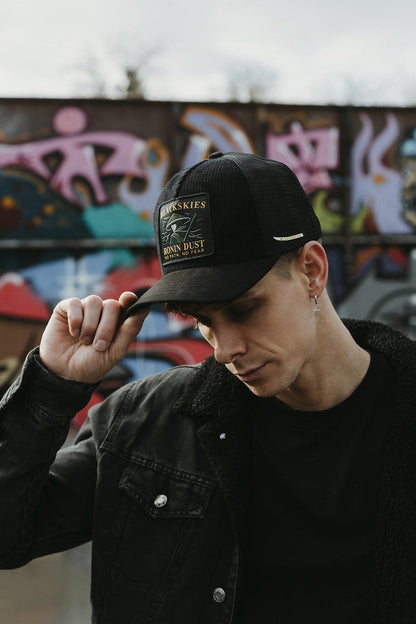 Person wearing a black cap with a logo, standing in front of a graffiti wall.